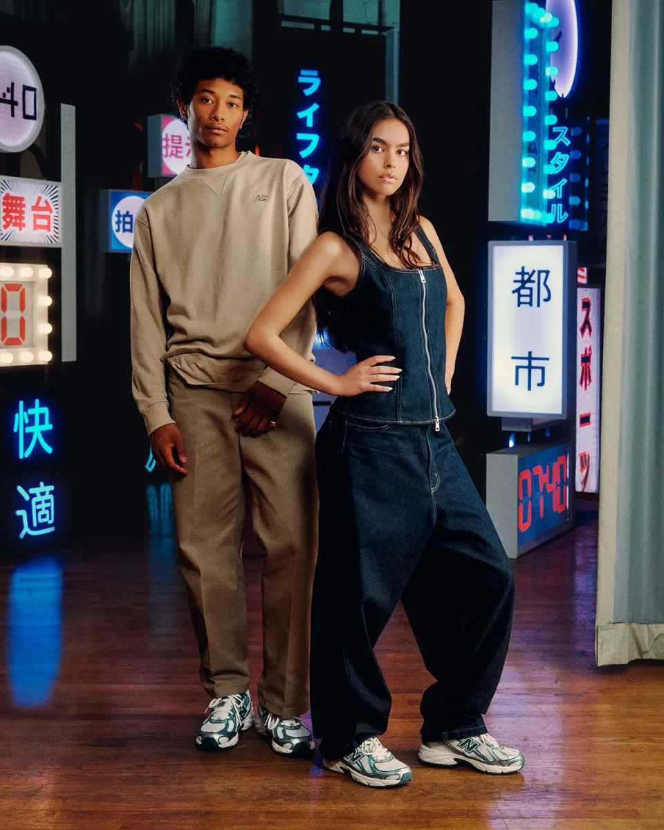 A boy and a girl wear the New Balance 740 on their feet. The girl is wearing a denim two-piece, the boy a brown jumper with brown trousers. They are standing in front of LED signs with Chinese characters.